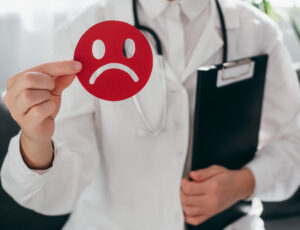 Woman doctor in white uniform with stethoscope holding little red angry emoticon and clipboard. Emotional intelligence, balance emotion control, mental health assessment, bipolar disorder concept